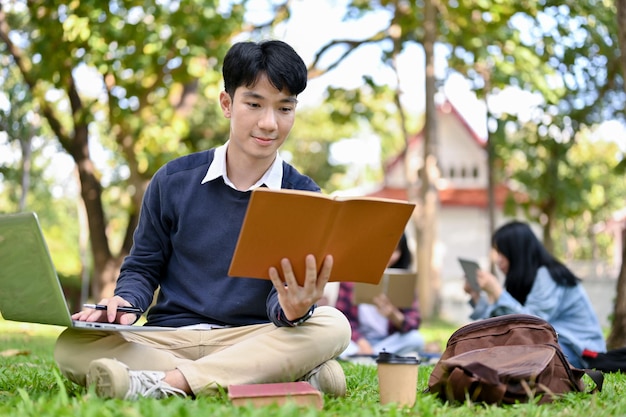 Smart Asian male student reading a book using laptop doing homework in the campus's park