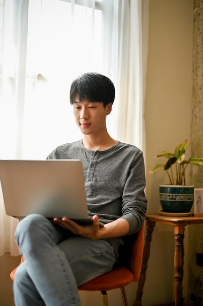 Smart Asian man focusing on his work on laptop using laptop on a chair in his living room