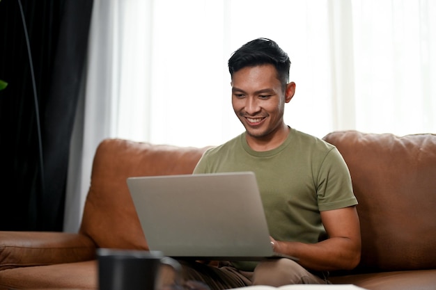 Smart Asian man using laptop on a sofa in his living room work from home free time leisure