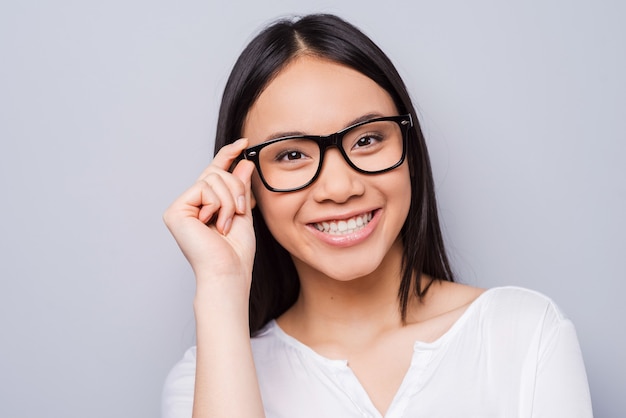 Smart and beautiful. Beautiful young Asian woman adjusting her eyewear and looking at camera with smile while standing against grey background