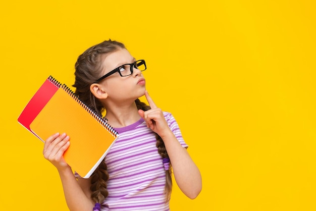 A smart beautiful girl with pigtails on an isolated background holds notebooks in her hand A schoolgirl with books and glasses on a yellow background