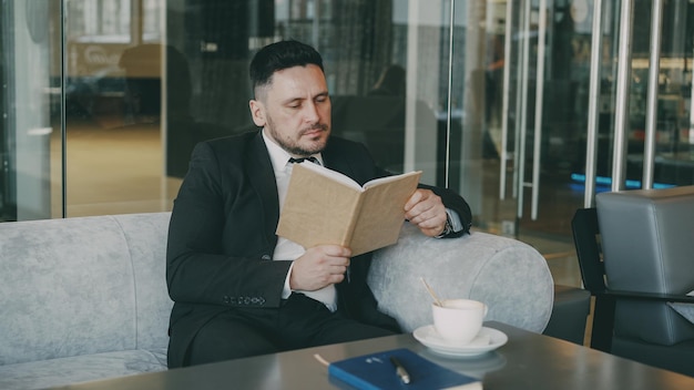 Smart Caucasian businessman in formal clothes reading text book with and drinking coffee in cafe