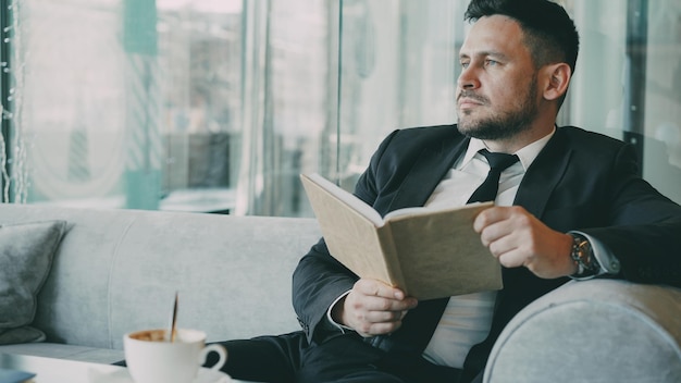 Smart Caucasian businessman in formal clothes reading text book with and drinking coffee in classy cafe at lunch break