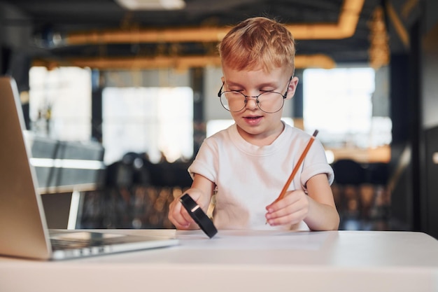 Smart child in casual clothes with laptop on table have fun with magnifying glass