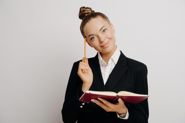 Smart female worker generates business ideas with red notebook pencil and smile against light wall