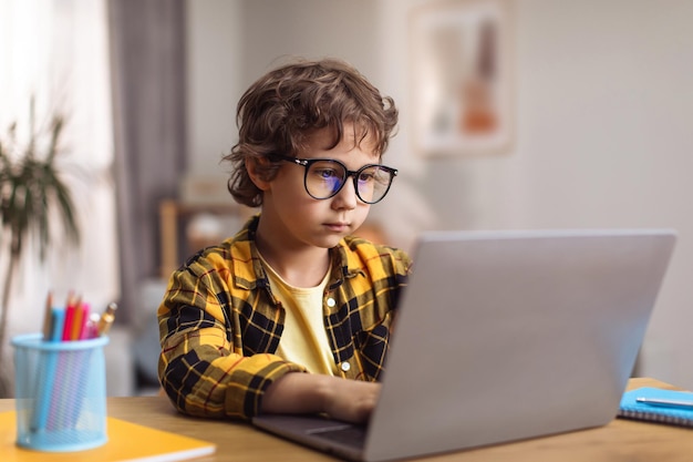 Smart generation Concentrated little boy in eyeglasses typing on laptop at home creating new game online free space