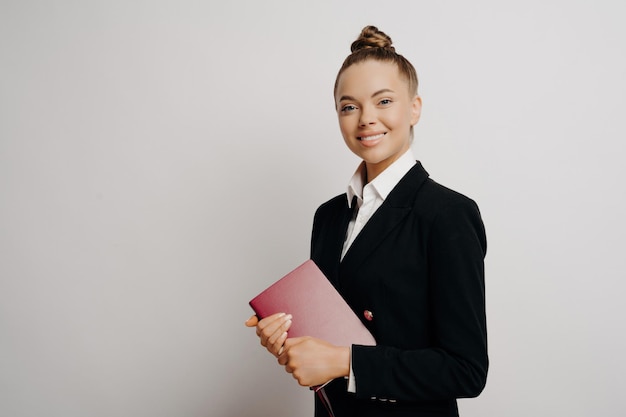 Smart happy female business person holds red notebook smiles at camera posing isolated against light background