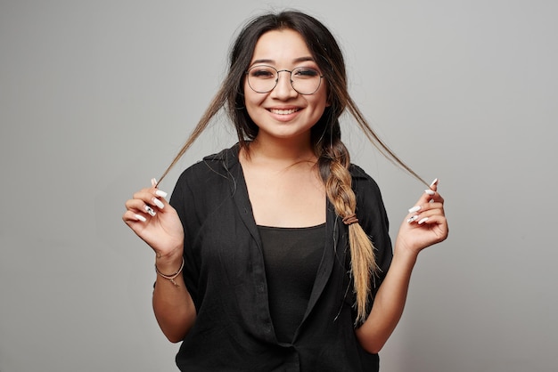 Smart Kazakh Asian student girl with pigtail and glasses touches hair posing in black clothes smiling on a white background