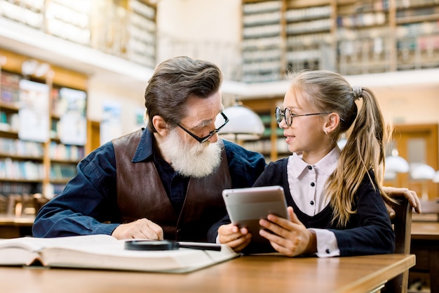 Smart kid girl sitting at the table in old library and holding tablet, showing something on tablet to her teacher or grandfather. Senior man with his student little girl in library