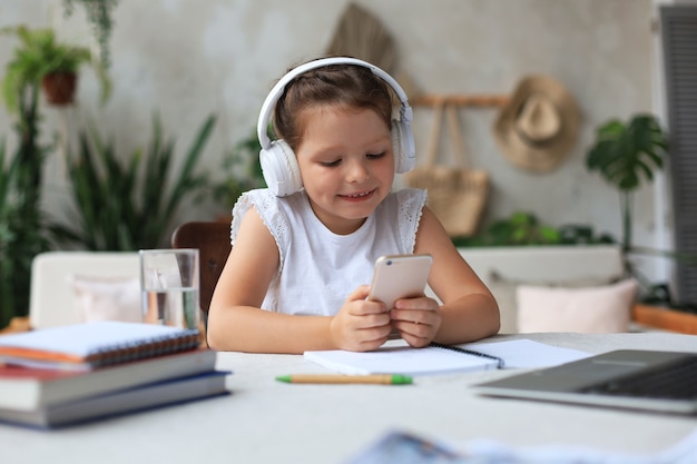 Smart little girl in headphones do homework online class on smartphone, small child in earphones study on Internet, have web conference or lesson during quarantine.