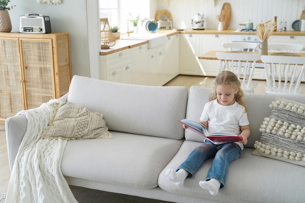 Smart little kid girl reading story sitting on cozy couch alone at home, preschool child reads fairytale, holding book, studying literature for children in living room. Children's education.