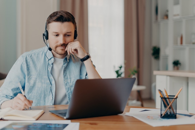 Smart man freelancer listens important info online makes necessary notes in diary works on laptop at home poses at desktop wears denim shirt poses over blurred background. Hotline support service