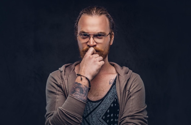 Smart pensive redhead hipster with full beard and glasses dressed in casual clothes, poses with hand on chin in a studio. Isolated on the dark background.