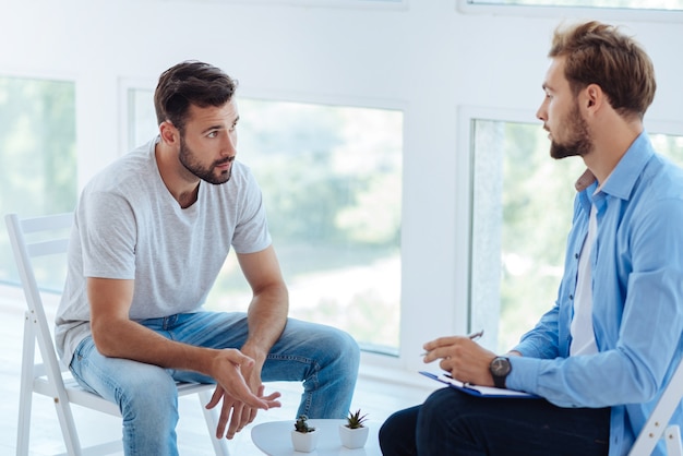 Smart professional male therapist looking at his patient and taking notes while listening about his problems