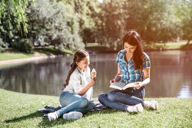 Smart woman is sitting at the edge of small lake with her daughter and reading a book. Her kid is sitting besides her and eating ice cream. Girl is looking at what mom is doing right now.