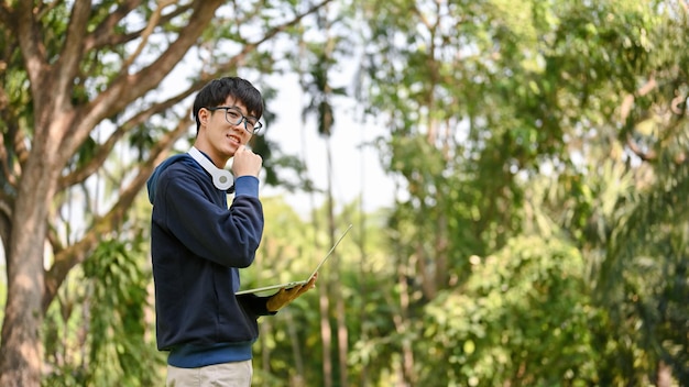 Smart young Asian male college student with his laptop standing in the greenery park