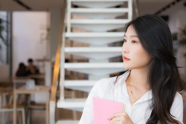 Smart young business woman wearing white shirt and blue jean holding pink book standing in front of stairway in conceptual of business successful