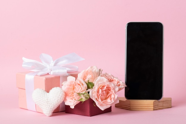 Smartphone on the background of a pink gift box with roses and a knitted white heart on a light pink background with a place for text