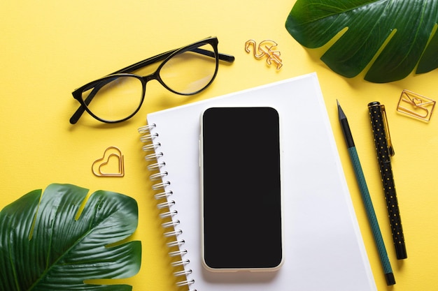 Smartphone blank notepad glasses pen paper clips pencil on a yellow background Blogging in the workplace is a business Copy space Flat lay top view