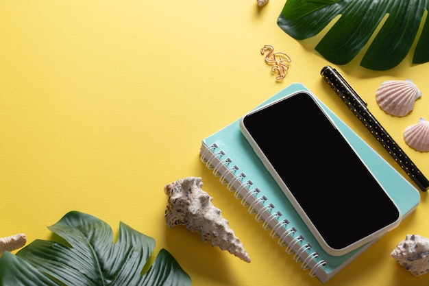 A smartphone glasses a blank notebook and Monstera Deliciosa leaves and shells on a yellow background Blogging in the summer at the workplace is a business