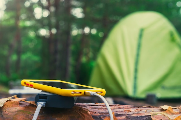 Smartphone is charged using a portable charger. Power Bank charges the phone outdoors with a backpack against the backdrop of a tent.