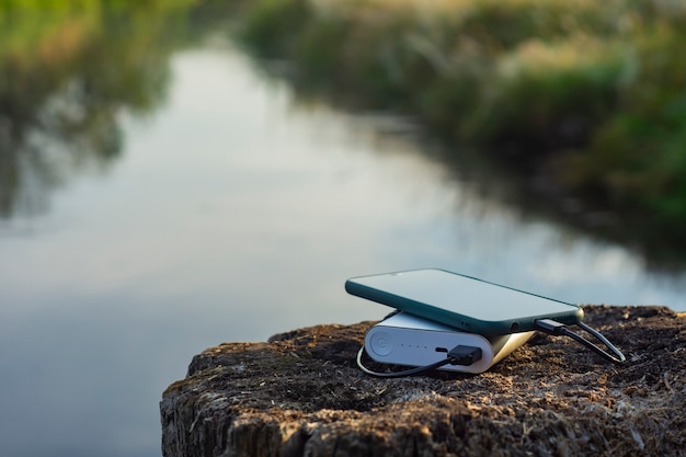 Smartphone is charged using a portable charger. Power Bank charges the phone outdoors with a backpack for tourism in the background of nature and the river.