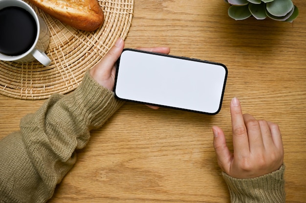 A smartphone mockup in a horizontal position is in a woman's hand over wood tabletop top view