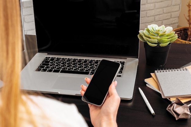 Smartphone with black screen in female hands. Computer, keyboard and office supplies on a background