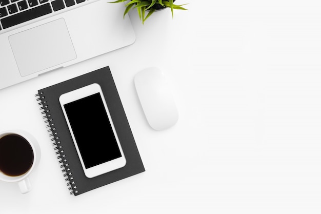 Smartphone with blank screen is on top of white office desk table with laptop and supplies.