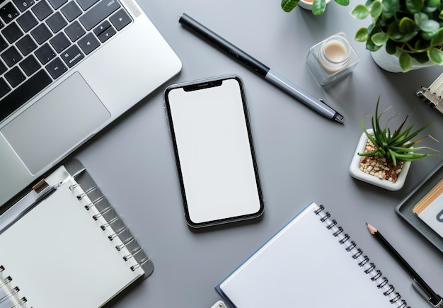 A smartphone with a blank white screen on a work desk surrounded by a laptop notepad and stationery