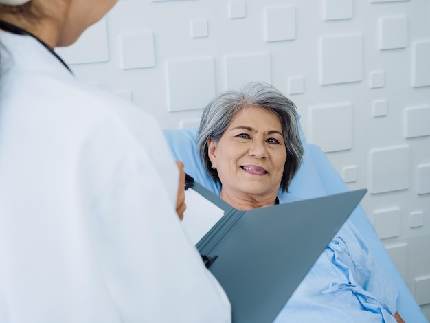 Smile Asian senior woman grey hair patient lying on bed recovering while young female doctors visit explaining the symptoms and examination notes and medical healthcare documents in hospital room