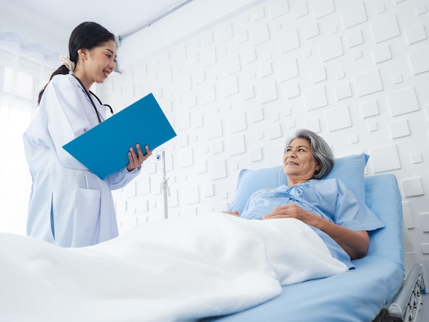 Smile Asian senior woman grey hair patient lying on bed recovering while young female doctors visit explaining the symptoms and examination notes and medical healthcare documents in hospital room