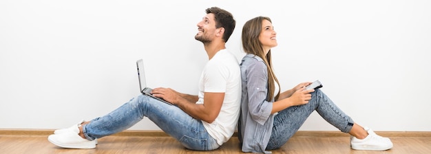 The smile couple sit with a laptop and a tablet near the empty wall