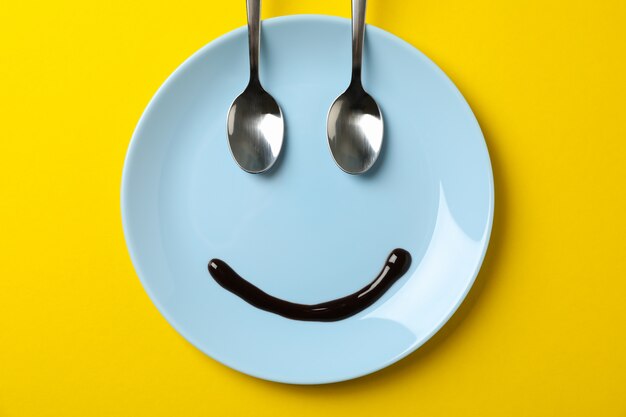 Smile face made of plate, chocolate syrup and spoons on yellow background, top view