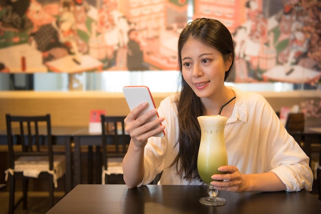 smile friendly female traveler holding japan ice matcha green tea milk and using mobile phone chatting with friends in leisure afternoon sitting on japanese drink restaurant.