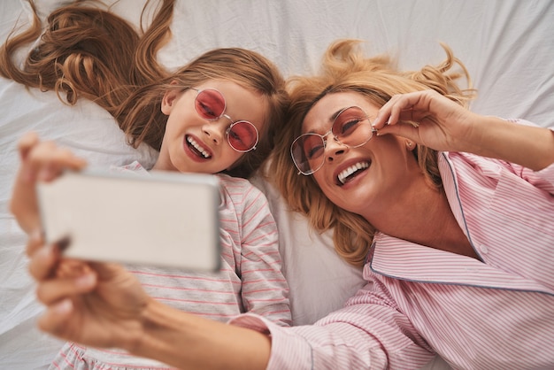 Smile! Top view of young beautiful mother and her cute daughter taking selfie with smart phone and smiling while lying on the bed at home