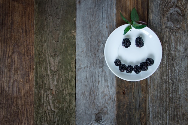 Smiley shape smile from blackberry berries on a white plate on a wooden table