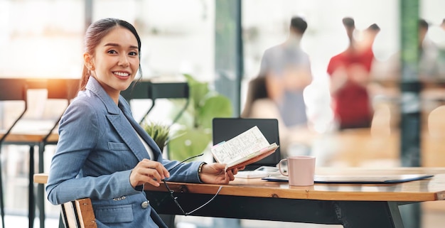 Smiley young businesswoman in suit holding business plan note and sitting at desk in modern coworking office