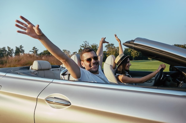 Smilinbg couple driving cabriolet and raising hands together