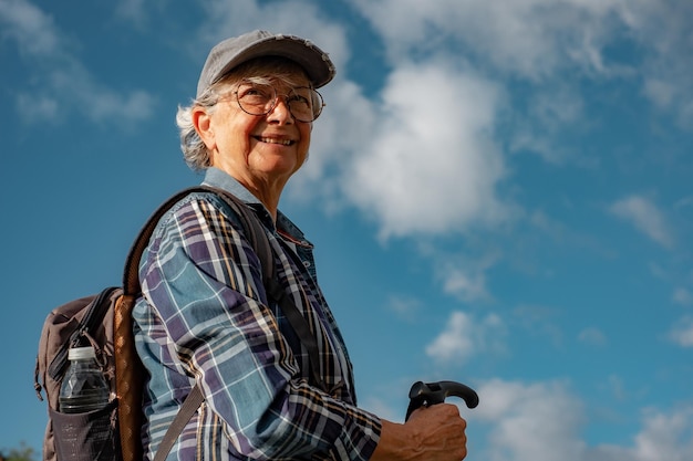 Smiling active senior woman enjoying healthy lifestyle goes trekking in mountain environment Elderly caucasian female with hat and backpack walking with sport stick