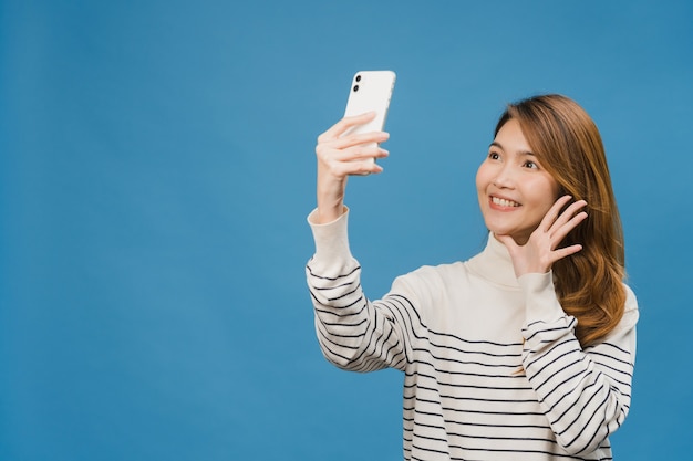 Smiling adorable Asian female making selfie photo on smart phone with positive expression in casual clothing and stand isolated on blue wall