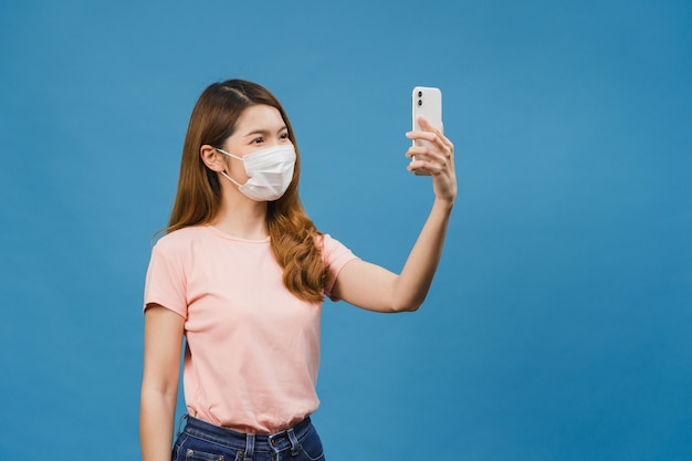 Smiling adorable Asian female wearing medical face mask making selfie photo on smart phone with positive expression in casual clothing and stand isolated on blue wall