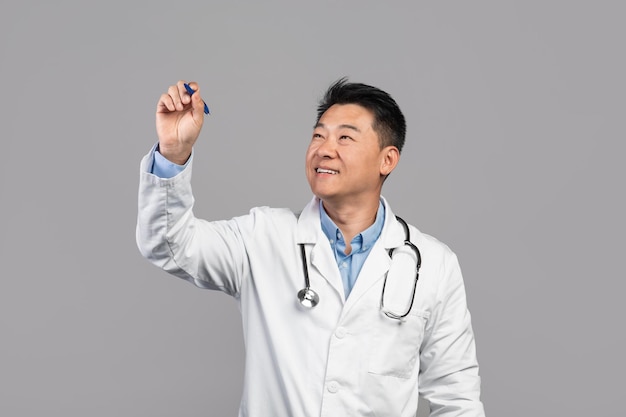 Smiling adult asian guy doctor in white coat with stethoscope writes on virtual blackboard with blank space