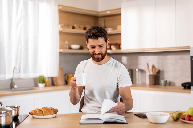 Smiling adult caucasian man in white tshirt enjoy cup of coffee free time reading book at table