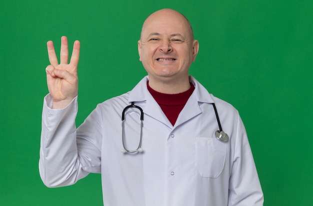 Smiling adult slavic man in doctor uniform with stethoscope gesturing three with fingers