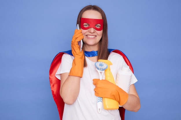 Smiling adult woman housewife wearing gloves and superhero costume holding detergent isolated over blue background talking on mobile phone with cheerful face