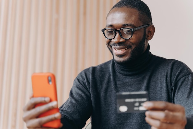 Smiling African American businessman using credit card and smartphone while sitting at workplace