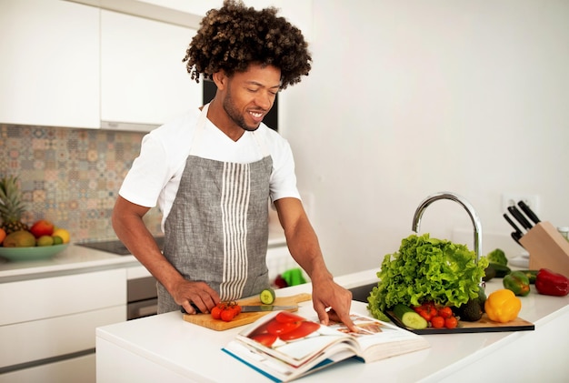 Smiling african american guy reading paper culinary book in kitchen