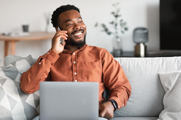 Smiling African American Guy Using Laptop Talking On Phone Indoors