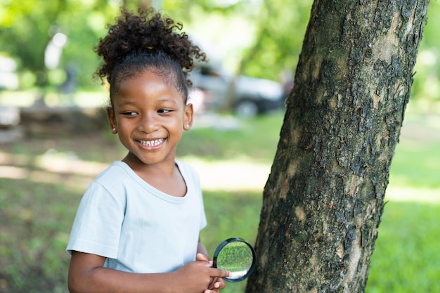 Smiling African American little girl holding magnifying glass to explore on tree in park